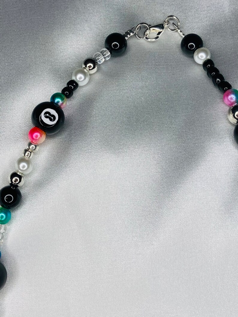 May include: A beaded necklace with a black 8-ball charm, black, white, silver, and iridescent beads. The necklace has a silver clasp and is displayed on a white satin background. The beads are various sizes and shapes.