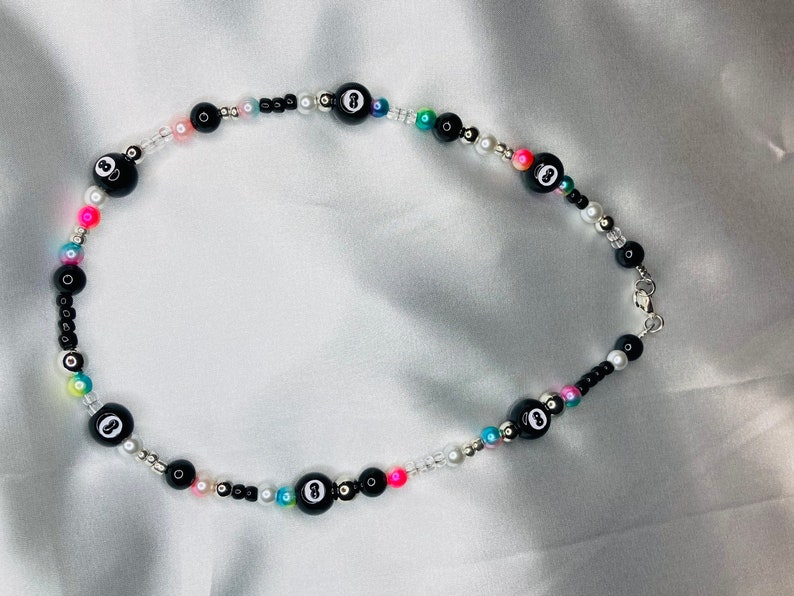 May include: A necklace with black eight ball beads, small black, white, and colourful beads. The necklace has a silver clasp. The beads are measured in centimetres. The necklace is displayed on a white surface.