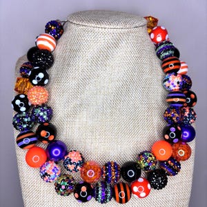 May include: A multi-strand chunky beaded necklace featuring a vibrant assortment of spherical beads in orange, black, purple, and white. The beads include solid colors, glittery textures, striped patterns, and polka dot designs arranged in a layered style.