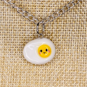 May include: A silver chain necklace with a white ceramic fried egg charm. The egg has a yellow yolk with a smiling face.