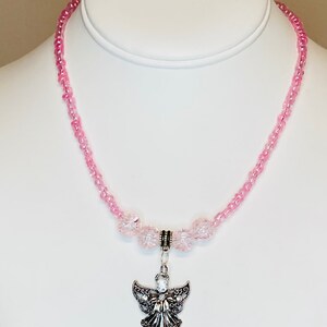 May include: A pink beaded necklace with a silver angel pendant. The necklace features small, light pink beads and larger, clear pink beads near the pendant. The angel pendant has detailed wings and a central crystal.