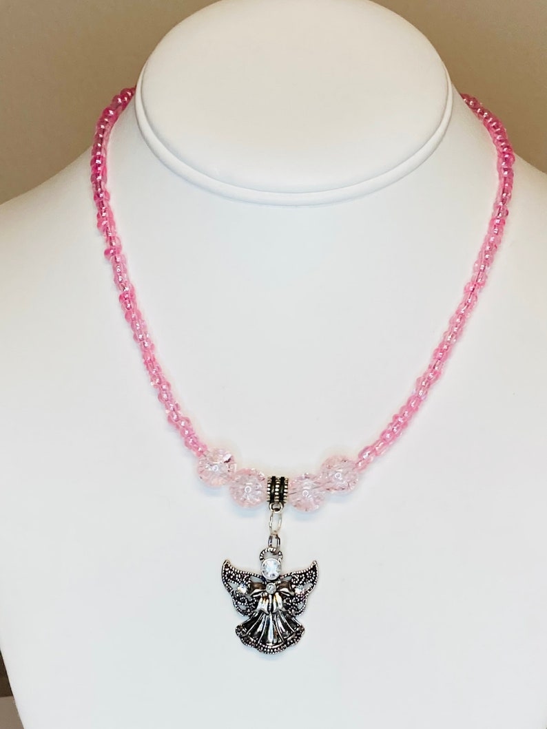 May include: A pink beaded necklace with a silver angel pendant. The necklace features small, light pink beads and larger, clear pink beads near the pendant. The angel pendant has detailed wings and a central crystal.