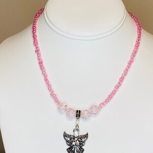 May include: A pink beaded necklace with a silver angel pendant. The necklace features small, round pink beads and larger, faceted pink beads. The angel pendant is detailed with intricate wings and a central gemstone.