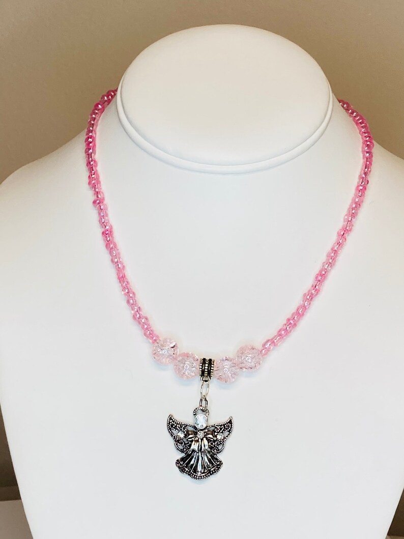 May include: A pink beaded necklace with a silver angel pendant. The necklace features small, round pink beads and larger, faceted pink beads. The angel pendant is detailed with intricate wings and a central gemstone.