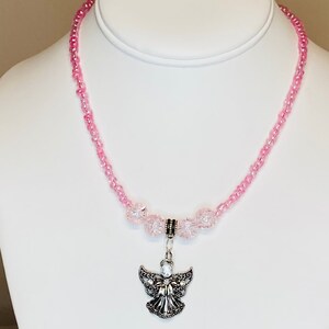 May include: A pink beaded necklace with a silver angel pendant. The necklace features small pink beads and larger, clear pink beads near the pendant. The angel pendant has detailed wings and a small gem.