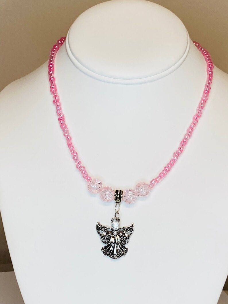 May include: A pink beaded necklace with a silver angel pendant. The necklace features small pink beads and larger, clear pink beads near the pendant. The angel pendant has detailed wings and a small gem.
