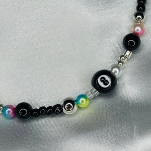 May include: A beaded necklace with a black eight ball bead, black beads, silver beads, and iridescent beads in pink, blue, and yellow. The necklace is shown against a silver satin backdrop.