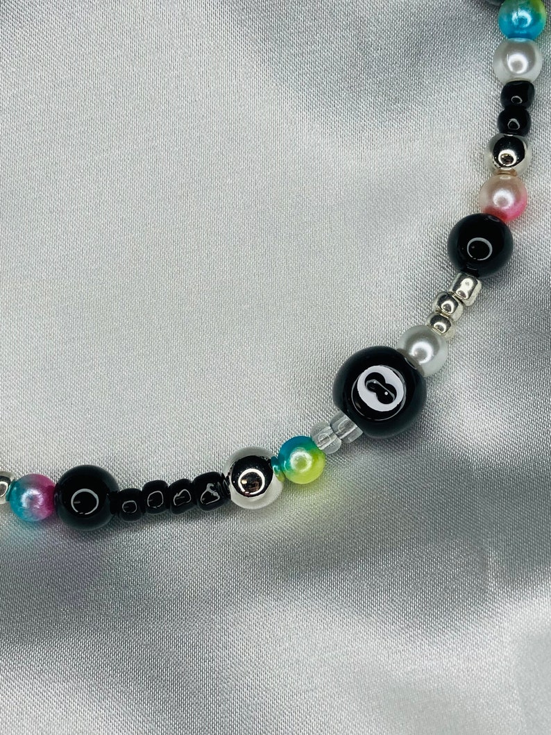 May include: A beaded necklace with a black eight ball bead, black beads, silver beads, and iridescent beads in pink, blue, and yellow. The necklace is shown against a silver satin backdrop.