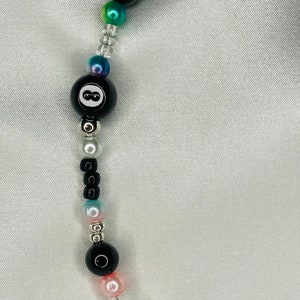 May include: A beaded necklace with a black 8-ball bead, iridescent beads, and clear seed beads. The necklace includes black, pink, and silver beads. The beads are strung on a clear cord.