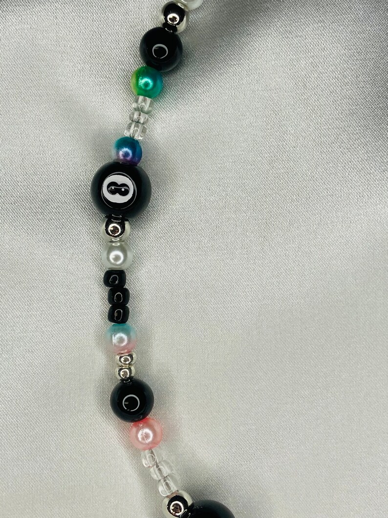 May include: A beaded necklace with a black 8-ball bead, iridescent beads, and clear seed beads. The necklace includes black, pink, and silver beads. The beads are strung on a clear cord.