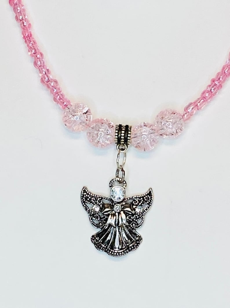 May include: A pink beaded necklace with a silver angel pendant. The angel pendant has a rhinestone in the center and is detailed with intricate wing and dress designs. The necklace features small pink beads and larger, textured pink beads.