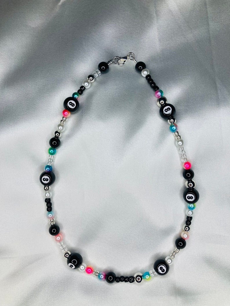 May include: A beaded necklace with black 8-ball beads, small black, white, and iridescent beads. The necklace has a silver clasp. The background is white.