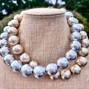 May include: A multi-strand necklace featuring large, round beads with a textured, metallic finish. The beads alternate between a warm gold tone and a cool silver tone, creating a reflective, crinkled surface appearance throughout the piece.