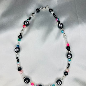 May include: A beaded necklace with black 8-ball beads, colourful and white beads. The necklace has a silver clasp. The necklace is displayed on a white satin background.