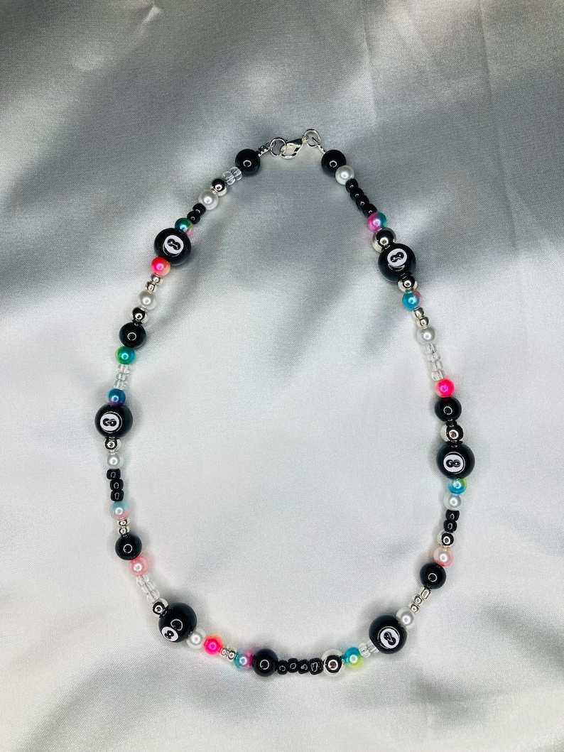 May include: A beaded necklace with black 8-ball beads, colourful and white beads. The necklace has a silver clasp. The necklace is displayed on a white satin background.