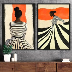 May include: Two framed art prints featuring black and white illustrations of women wearing striped dresses against a red background. The prints are hung on a grey wall above a wooden cabinet.