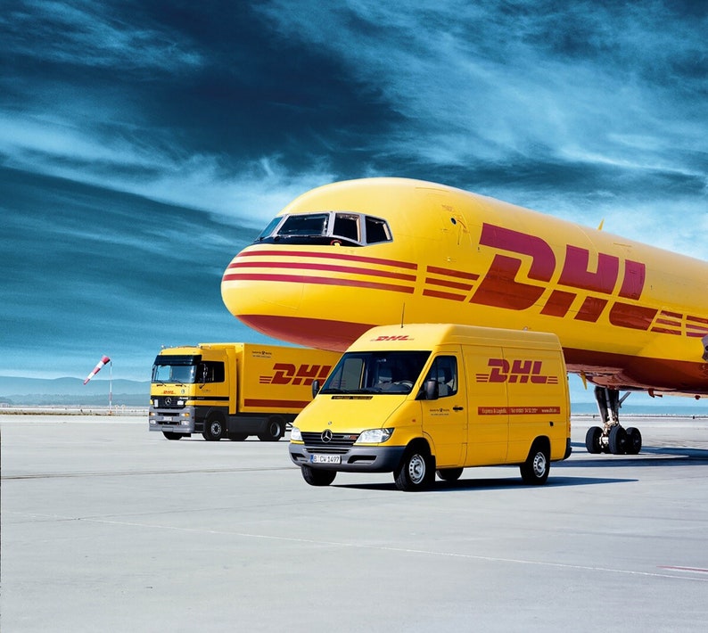 May include: A yellow DHL airplane, truck, and van are parked on a concrete surface. The vehicles and aircraft have the DHL logo in red. The sky is blue with white clouds. The airplane is the largest vehicle in the image.
