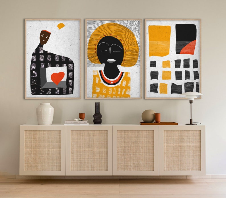May include: Three framed abstract art prints. The artwork features figures and shapes in black, orange, and white. The prints are displayed above a white cabinet with woven panels. A lamp and decorative items are on the cabinet.