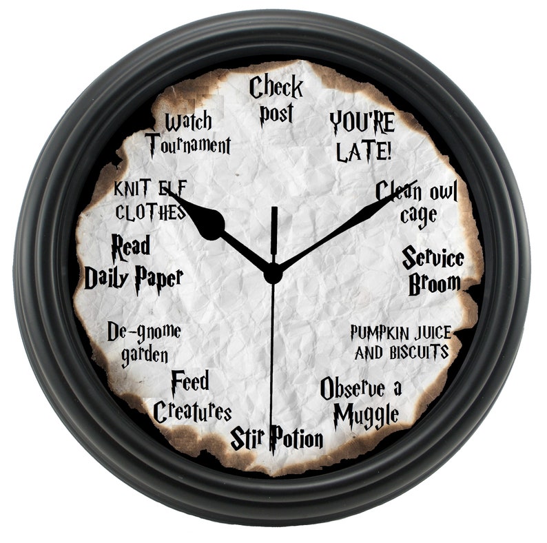 May include: A black framed clock with a white face and a burnt parchment paper design. The clock face has text around the edge, including "Watch Tournament", "Knit Elf Clothes", "Read Daily Paper", "De-gnome Garden", "Feed Creatures", "Stir Potion", "Check Post", "You're Late", "Clean Owl Cage", "Service Broom", "Pumpkin Juice and Biscuits", and "Observe a Muggle".