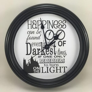 May include: Black clock with a white face and black hands. The clock face has the quote "Happiness can be found even in the darkest of times, if one only remembers to turn on the light." The quote is in a black font. The clock has a black frame with a silver rim.