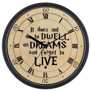 May include: Black framed clock with Roman numeral markings and a quote that reads "It does not do to dwell on dreams and forget to live".