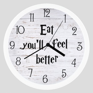 May include: White clock with black hands and black numbers on a white woodgrain background. The clock face has the text "Eat you'll feel better" in black.