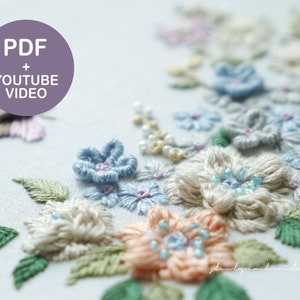 May include: Close-up of a floral embroidery design featuring pastel-colored flowers and green leaves. The flowers are in shades of blue, peach, and cream, with delicate bead accents. The text "PDF + YOUTUBE VIDEO" is in a purple circle.