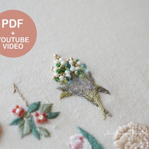 以下が含まれることがあります： クリーム色の生地に刺繍された花のクローズアップ。刺繍は、緑の葉、赤いベリー、ビーズのアクセントが付いたさまざまな花を特徴としています。画像には、ピンクの円の中に「PDF + YOUTUBE VIDEO」というテキストが含まれています。