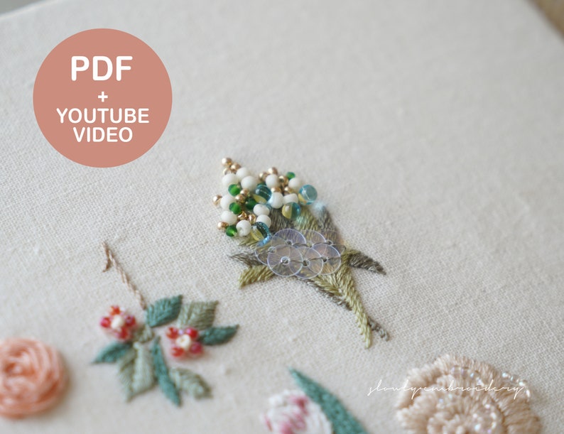 以下が含まれることがあります： クリーム色の生地に刺繍された花のクローズアップ。刺繍は、緑の葉、赤いベリー、ビーズのアクセントが付いたさまざまな花を特徴としています。画像には、ピンクの円の中に「PDF + YOUTUBE VIDEO」というテキストが含まれています。