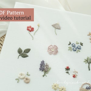 May include: A collection of embroidered floral designs, including red, blue, and pink flowers, and green leaves, on a white fabric background. The text "PDF Pattern + video tutorial" is in a pink box in the upper left corner. The embroidery is delicate and detailed.