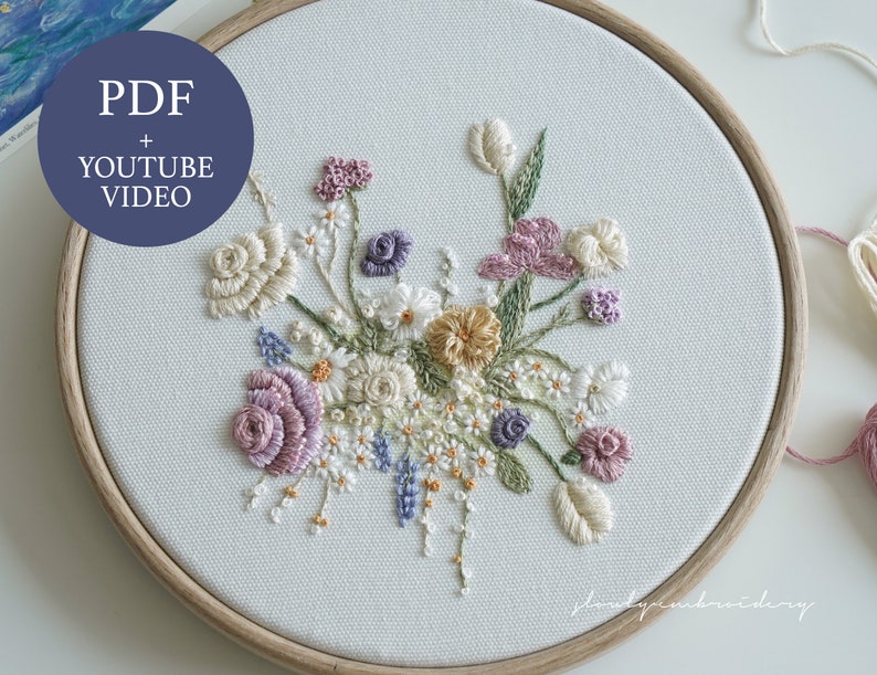 May include: Hand-embroidered floral design in a wooden hoop. The embroidery features a variety of flowers in shades of cream, pink, purple, and yellow, with green leaves. The text "PDF + YOUTUBE VIDEO" is in a blue circle. The design is on a light-colored fabric.