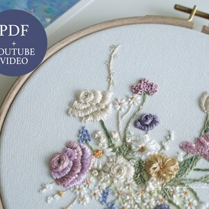 May include: A detailed floral embroidery design featuring a variety of flowers in shades of white, cream, lavender, and yellow. The embroidery is set in a wooden hoop and includes the text "PDF + YOUTUBE VIDEO".