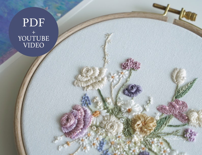 May include: A detailed floral embroidery design featuring a variety of flowers in shades of white, cream, lavender, and yellow. The embroidery is set in a wooden hoop and includes the text "PDF + YOUTUBE VIDEO".