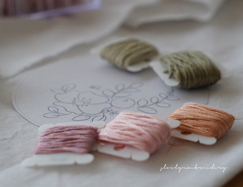 Puede incluir: Primer plano de suministros de bordado. Varios carretes de hilo en tonos rosa, melocot&oacute;n y verde est&aacute;n dispuestos sobre una superficie blanca. Un patr&oacute;n de bordado floral es visible, junto con el texto "slowlyembroidery".