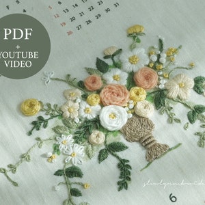 以下が含まれることがあります： 白い、桃色、黄色の花と緑の葉のブーケが特徴の刺繍された花の模様。刺繍は白い布に施されています。円内のテキストは「PDF + YOUTUBE VIDEO」と書かれています。