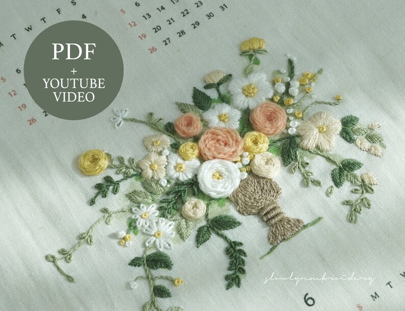 以下が含まれることがあります： 白い、桃色、黄色の花と緑の葉のブーケが特徴の刺繍された花の模様。刺繍は白い布に施されています。円内のテキストは「PDF + YOUTUBE VIDEO」と書かれています。
