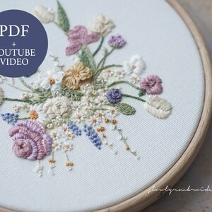 May include: An embroidered floral design in a wooden hoop. The embroidery features a variety of flowers in shades of cream, lavender, and peach, with green leaves and stems. The text "PDF + YOUTUBE VIDEO" is in a blue circle. The design is on a white fabric.