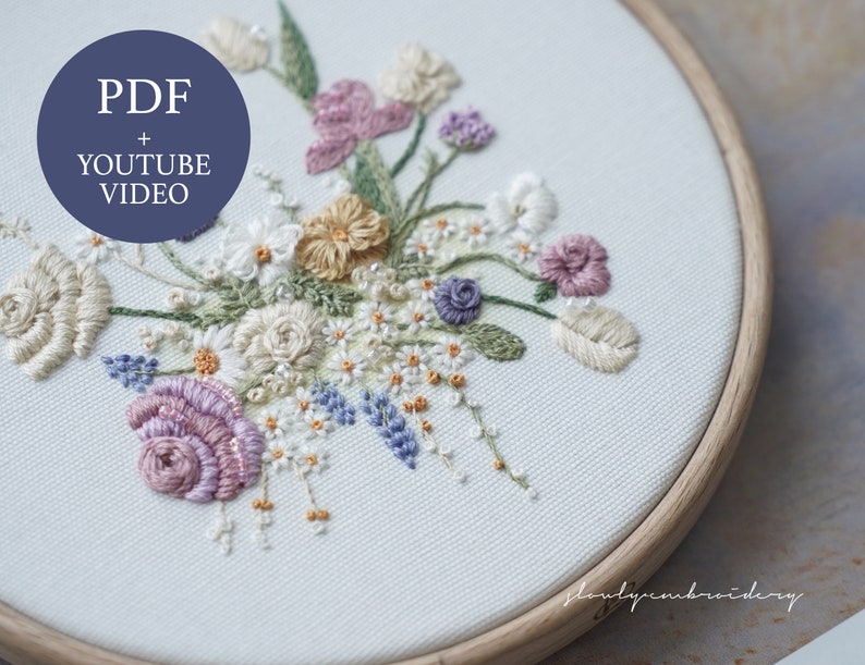 May include: An embroidered floral design in a wooden hoop. The embroidery features a variety of flowers in shades of cream, lavender, and peach, with green leaves and stems. The text "PDF + YOUTUBE VIDEO" is in a blue circle. The design is on a white fabric.