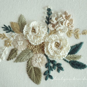 May include: Hand-stitched floral embroidery featuring cream-colored flowers, accented with green, beige, and gold thread. The detailed needlework showcases a variety of stitches, creating a textured, three-dimensional effect. The artwork is set against a white linen background.