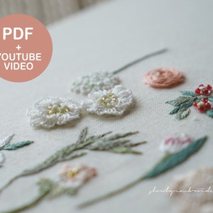 以下が含まれることがあります： 様々な手刺繍の花柄が施された白い布のクローズアップ。真珠のような中心を持つ白い花、ピンクとピーチ色の花、緑の葉が含まれています。画像には「PDF + YOUTUBE VIDEO」というテキストも含まれています。