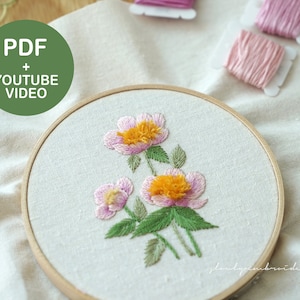 Op de afbeelding: Een borduurring met een bloemenontwerp met roze en gele bloemen en groene bladeren. De tekst "PDF + YOUTUBE VIDEO" staat in een groene cirkel. Borduurgaren en een schaar staan op de achtergrond.
