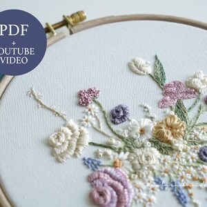 May include: An embroidery hoop with a floral design. The embroidery features a variety of flowers in shades of white, pink, purple, and yellow, with green leaves. The text "PDF + YOUTUBE VIDEO" is in a blue circle.