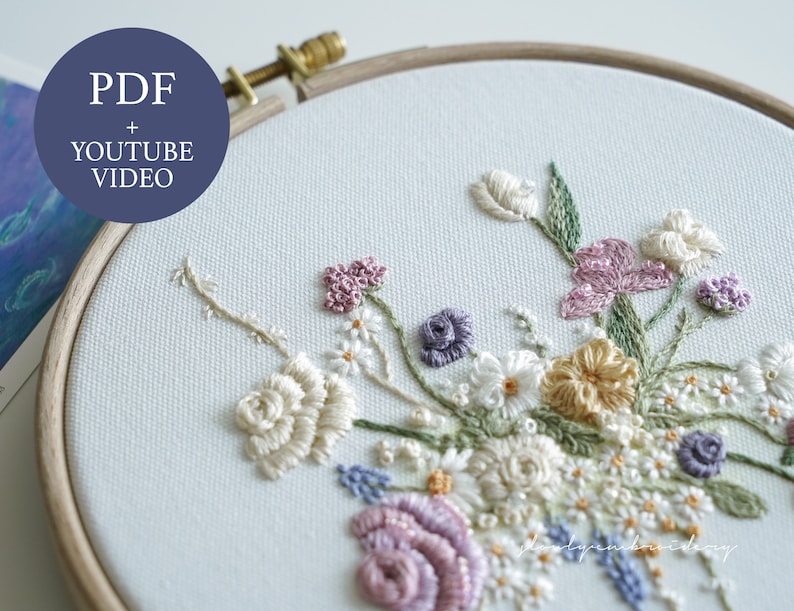 May include: An embroidery hoop with a floral design. The embroidery features a variety of flowers in shades of white, pink, purple, and yellow, with green leaves. The text "PDF + YOUTUBE VIDEO" is in a blue circle.