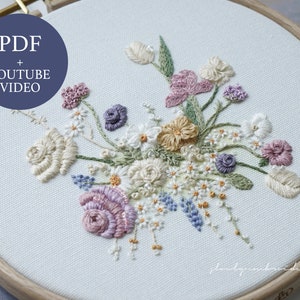 May include: Embroidered floral design in a wooden hoop. The artwork features a variety of flowers in shades of cream, pink, purple, and yellow, with green leaves. The text "PDF + YOUTUBE VIDEO" is in a blue circle. The embroidery is on a light blue fabric.