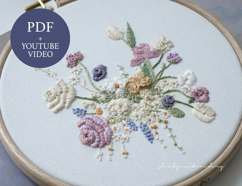 May include: Embroidered floral design in a wooden hoop. The artwork features a variety of flowers in shades of cream, pink, purple, and yellow, with green leaves. The text "PDF + YOUTUBE VIDEO" is in a blue circle. The embroidery is on a light blue fabric.