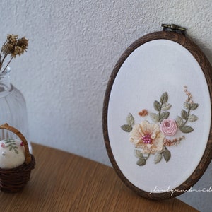 Puede incluir: Bastidor de bordado ovalado con dise&ntilde;o floral. El bordado presenta una gran flor color melocot&oacute;n con centro rosa, rodeada de rosas rosas y hojas verdes. El marco de madera tiene un acabado marr&oacute;n oscuro.