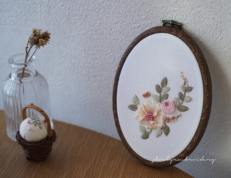 Puede incluir: Bastidor de bordado ovalado con dise&ntilde;o floral. El bordado presenta una gran flor color melocot&oacute;n con centro rosa, rodeada de rosas rosas y hojas verdes. El marco de madera tiene un acabado marr&oacute;n oscuro.