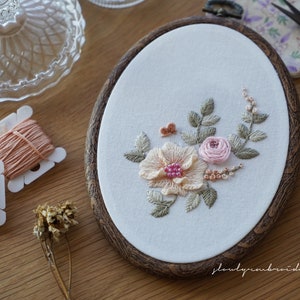 Puede incluir: Bastidor de bordado ovalado con un dise&ntilde;o floral que incluye una flor color melocot&oacute;n, una rosa rosa y hojas verdes. El bordado est&aacute; sobre tela blanca, enmarcado por un aro de madera oscura. Se ven carretes de hilo y tijeras.