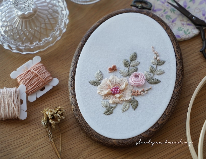 Puede incluir: Bastidor de bordado ovalado con un dise&ntilde;o floral que incluye una flor color melocot&oacute;n, una rosa rosa y hojas verdes. El bordado est&aacute; sobre tela blanca, enmarcado por un aro de madera oscura. Se ven carretes de hilo y tijeras.