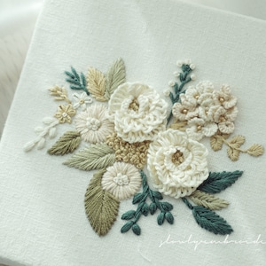 May include: A square, off-white canvas features an embroidered floral design. The embroidery includes white, cream, and tan flowers, with green and teal leaves. The design is detailed with small, gold-colored beads.